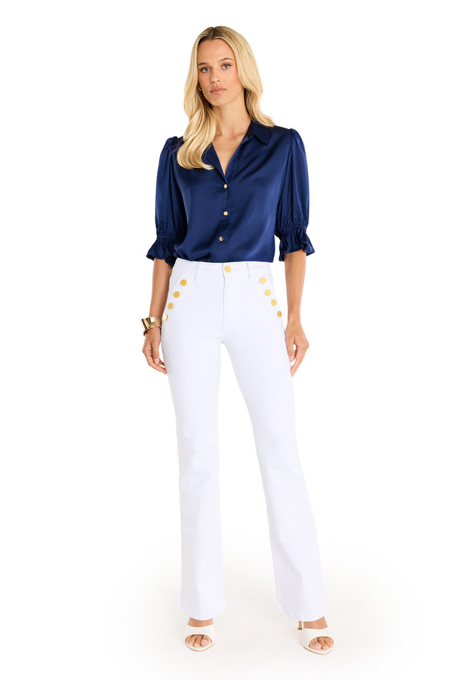 A woman faces forward in the Cindy Ruffle Sleeve Blouse—navy satin with subtle puffed sleeves and gold buttons—paired with high-waisted white pants featuring gold buttons. She has long blonde hair and wears open-toed heels.