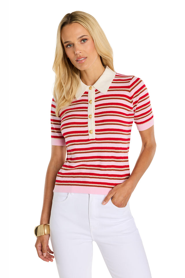 A woman with long blonde hair wears the Leya Polo Shirt – Summer Multi, featuring red, pink, and white stripes with a white collar and gold embossed buttons. She pairs it with white pants and a gold bracelet against a white background.