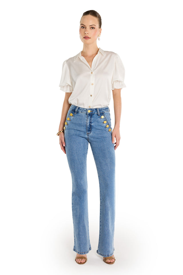 A woman stands against a white background wearing a white puff-sleeve blouse and Allegra Bootcut Jean - Medium Blue with gold detailing on the pockets. Her hair is tied back, and she completes the look with sandals.