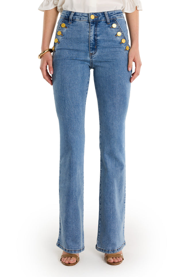 Someone wears the Allegra Bootcut Jean - Medium Blue, featuring a high waist and gold buttons in two vertical rows on each side. The bootcut jeans have a slight flare at the bottom, styled with sandals and a white top.
