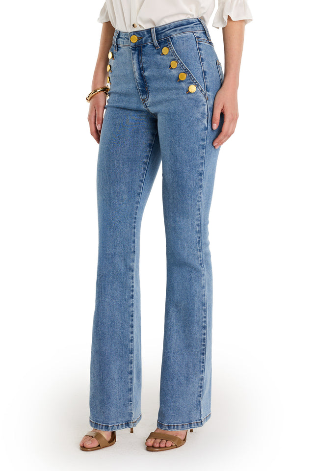 A person wears the Allegra Bootcut Jean - Medium Blue with gold hip detailing, a white blouse, tan heeled sandals, and a gold bracelet, standing against a white background.