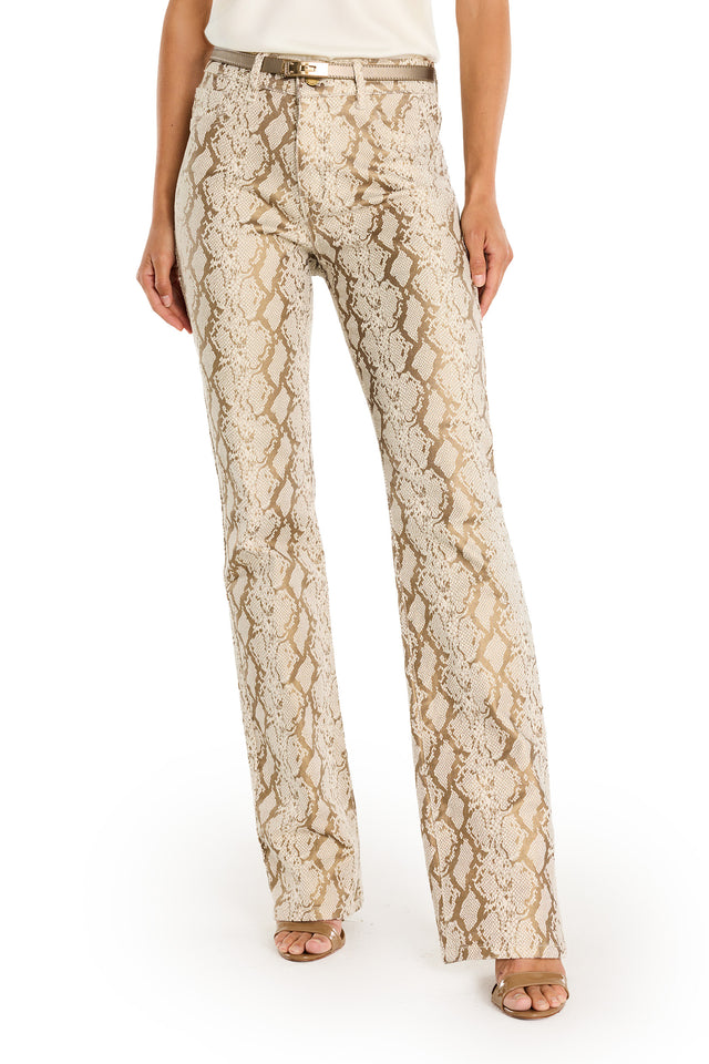 A person wearing the Aria Flare Jean - Gold Python, featuring a beige and gold snakeskin pattern, paired with a white sleeveless top, beige belt, and open-toe heels, stands against a white background.