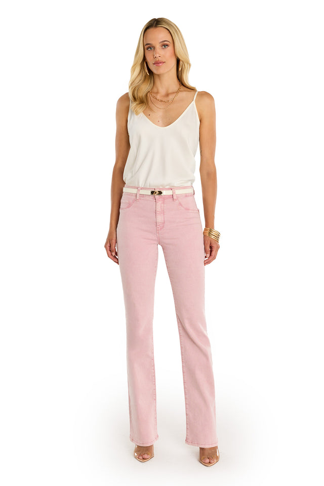 A woman with long blonde hair stands facing forward, wearing the Aria Flare Jean - Blush with a white sleeveless top and belt. She accessorizes with bracelets, necklaces, and nude heels against a white background.