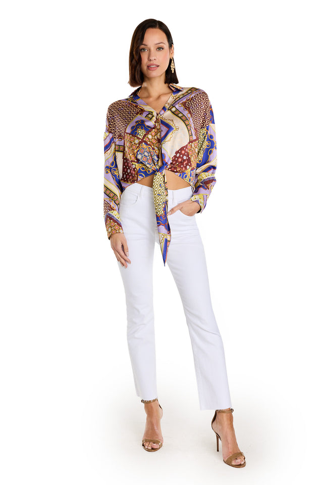 A woman with shoulder-length brown hair stands against a white background in the Cecilia Silk Tie-Front Blouse - Riviera Chain, paired with white pants, gold open-toe heels, earrings, and the blouse tied at the waist.