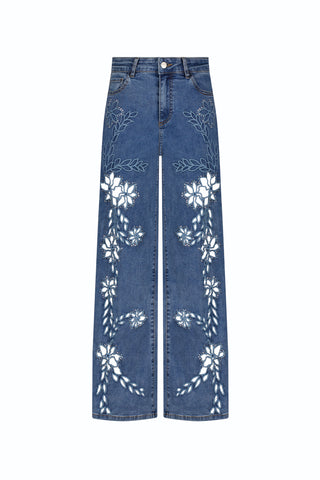 The Zenya Crystal High Rise Jean in Medium Blue (Preorder) features a wide-leg fit, four-way stretch denim, and white floral embroidery flowing symmetrically from thighs to ankles.