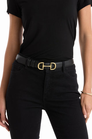 Wearing a black t-shirt and black pants, the person sports the Bettie Belt - Black with a gold horsebit buckle; their left hand is by their side, and a gold bangle adorns their right wrist.