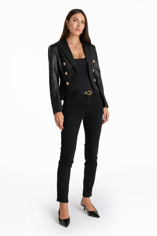 A woman stands against a white background wearing the Gabrielle Leather Double Breasted Blazer - Black with gold buttons, a black top, black skinny jeans with a belt, and black heels. The tailored blazer flatters her silhouette. She has long brown hair and a neutral expression.