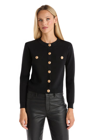 A woman with straight blonde hair wears the Loretta Knit Cardigan Top in black, featuring padded shoulders and gold dome buttons, paired with matching black leather pants against a white background.