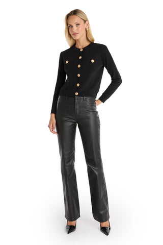 A woman with blonde hair stands confidently against a white background wearing the Loretta Knit Cardigan Top - Black, featuring padded shoulders and large gold dome buttons, styled with black leather pants and pointed-toe heels.