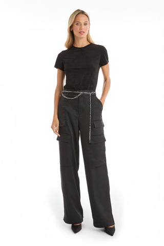 A woman with long blonde hair and a neutral expression stands against a white background wearing a black short-sleeve t-shirt, black wide-leg cargo pants with pockets, the Arden Chain Belt - Silver, and black pointed-toe heels.