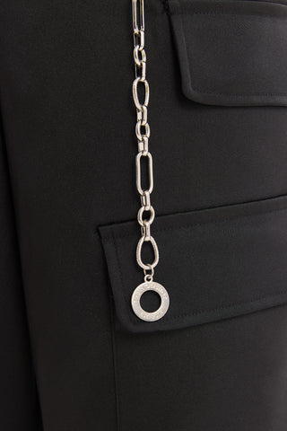 A close-up of the Arden Chain Belt - Silver, featuring a circular pendant attached to the pocket of a black garment. The smooth fabric and flap pocket complement the belt's elegant design.