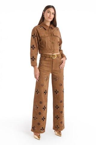 A woman stands confidently in Kaia Suede High Rise Jeans in cognac with floral cutouts, paired with a matching jacket, a belt featuring a gold buckle, and gold pointed-toe heels against a white background.