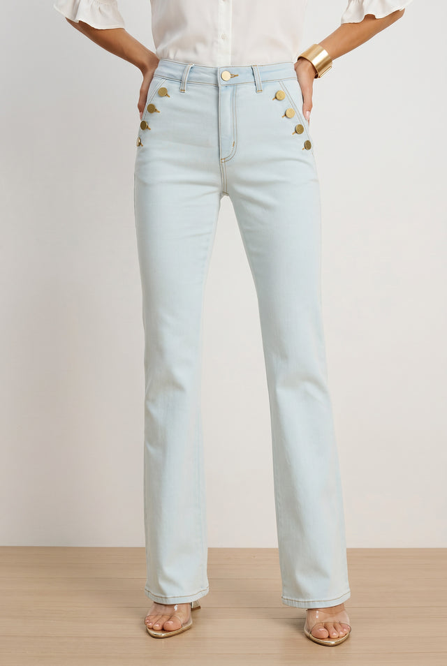 A person in Allegra Bootcut Jean - Cloud, a white blouse, gold bracelet, and open-toe heels stands on a light wooden floor. The high-rise jeans feature light-wash denim and gold button details at the front pockets.