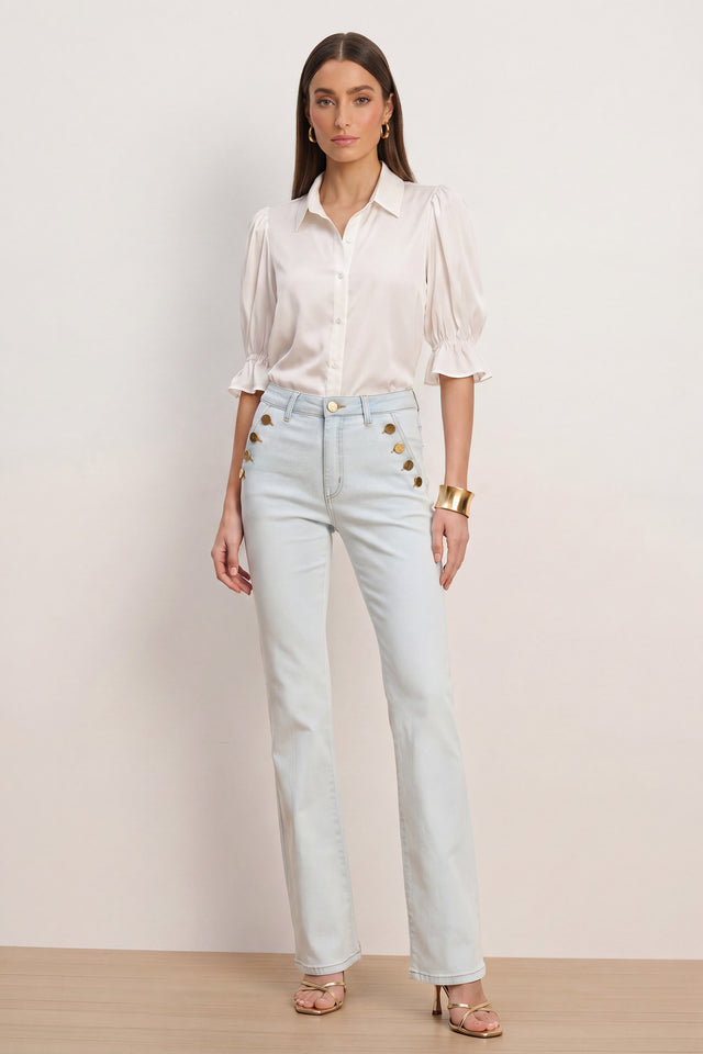 A woman stands against a plain background wearing a white blouse with puffed sleeves, Allegra Bootcut Jean - Cloud with gold buttons, gold jewelry, and nude heels.