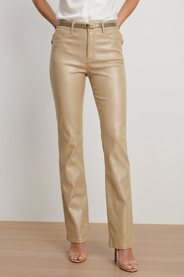 A person wearing Aria Coated Flare Jean in Champagne with a matching belt, a white top, and beige heeled sandals, standing on a wooden floor against a plain light background.