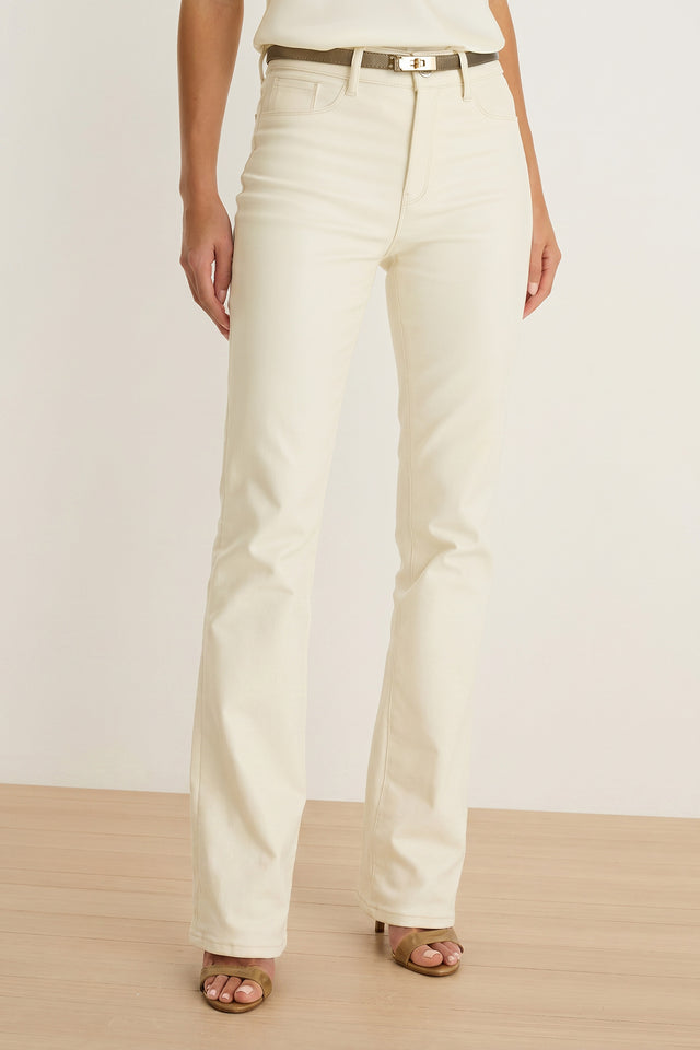 A person wearing Aria Coated Flare Jean in Vanilla, styled with a beige belt and brown open-toe heels, standing on a light wooden floor against a plain white background.