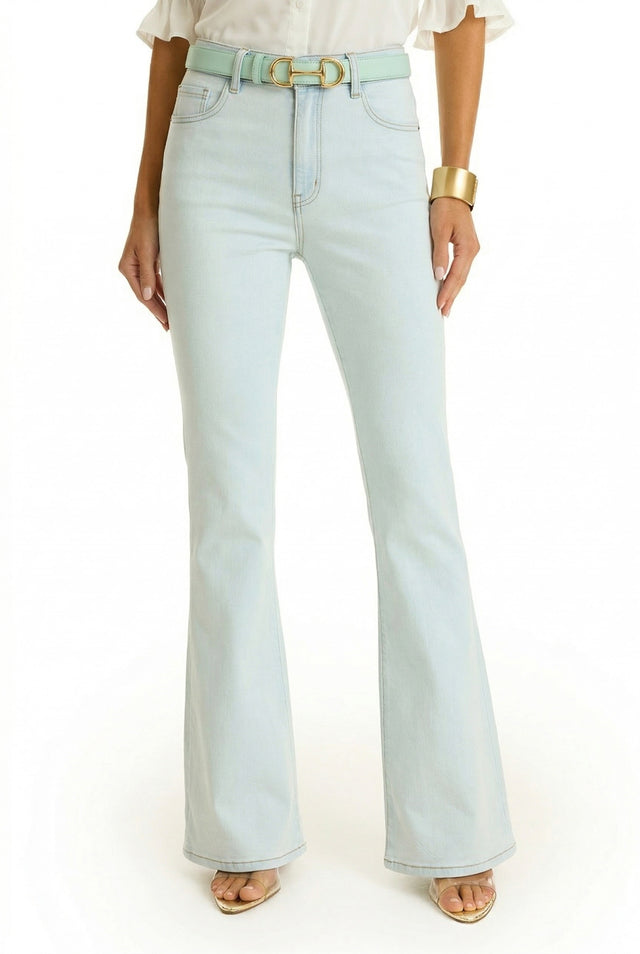 A person shown from shoulders down against a white background wears the Aria Flare Jean - Cloud, a high-rise, light-wash flared jean, paired with a light green belt, white blouse, gold bracelet, and open-toed heels.