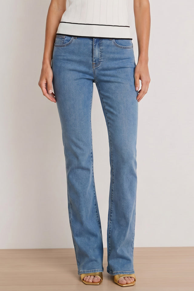 Model wears Aria Flare Jean - Medium Blue, paired with a white sleeveless ribbed top and tan open-toe heels, standing against a plain background with her hands at her sides.