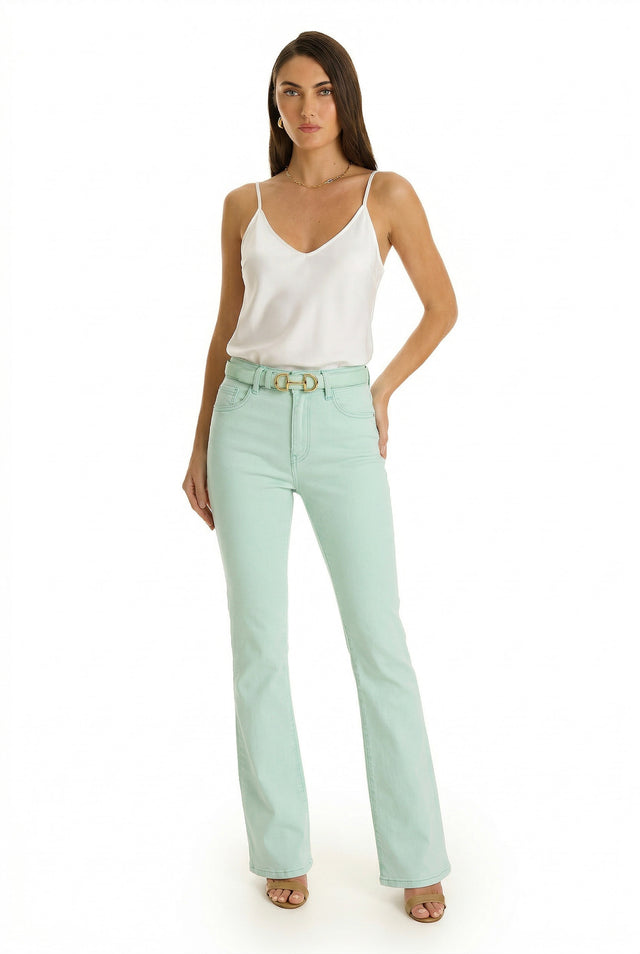 A woman with straight, long hair stands against a white background, wearing the Aria Flare Jean - Mint paired with a white sleeveless top, belt, and beige heeled sandals. One hand rests on her hip.