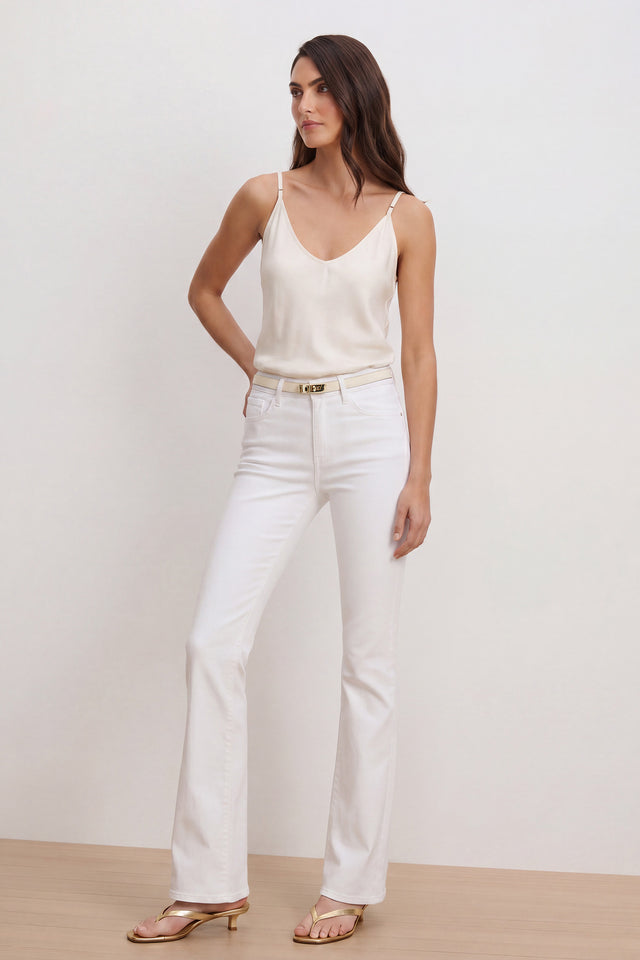 A woman poses confidently in a cream camisole, gold heeled sandals, and the Aria Flare Jean - White, featuring high-rise style and four-way stretch. She has long wavy brown hair and stands against a plain background.