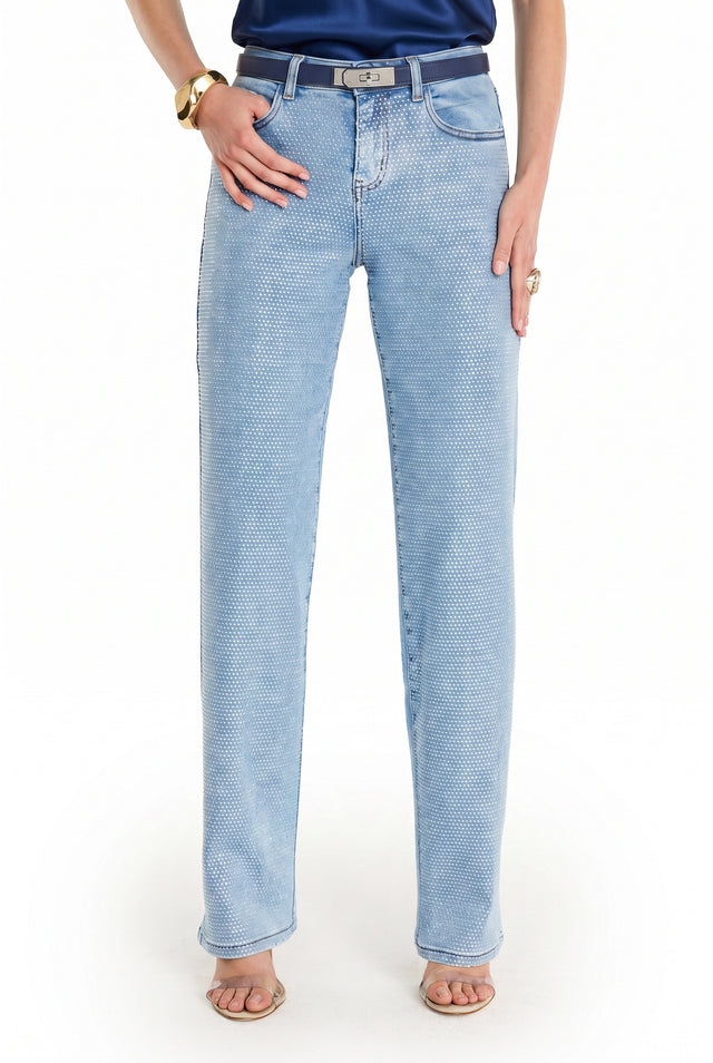 Wearing the Brynn Crystal Jean - Medium Blue with a belt, navy top, gold bracelet, and ring, a person stands against a white background. The high-rise wide-leg jeans offer stylish flair. The individual's head is not shown in the image.