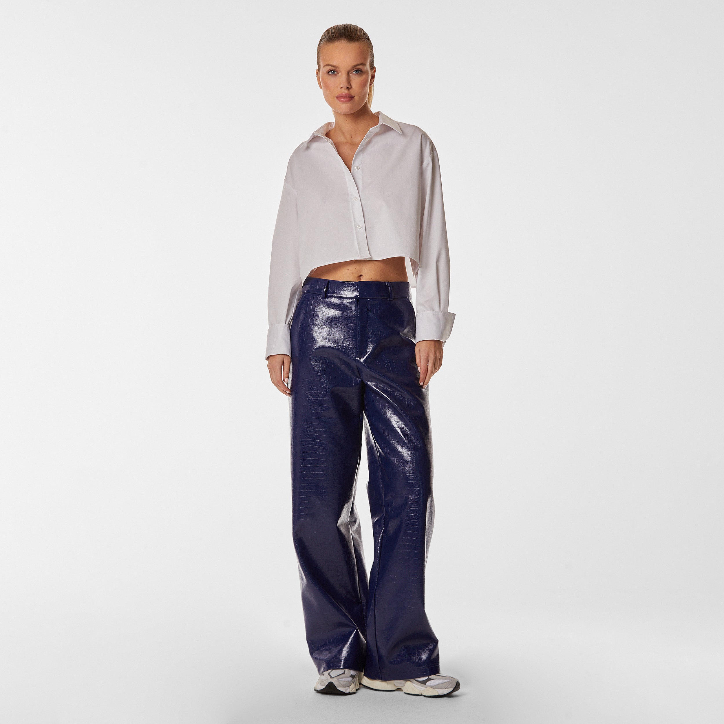 Full body front view of woman wearing white cropped button up shirt and navy blue faux leather croco pattern embossed wide leg pant.