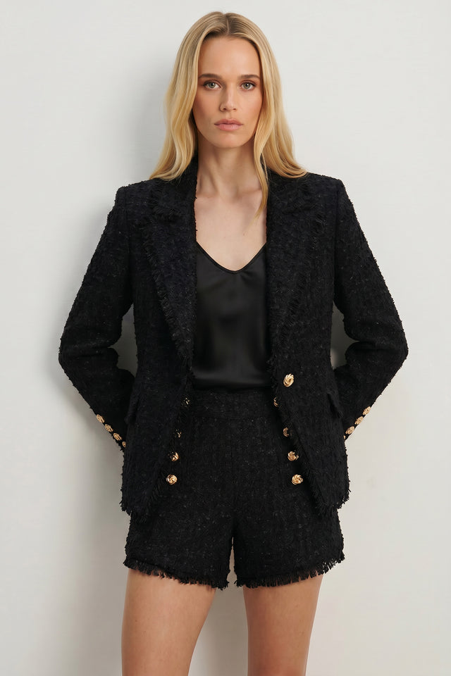 A woman with long blonde hair stands against a light background, wearing the Giselle Single Breasted Tweed Blazer - Black, styled with a black satin top and matching shorts. She looks confidently at the camera with her hands in her pockets.