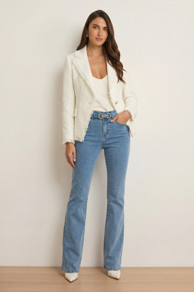 A woman stands against a plain background in the Giselle Single Breasted Tweed Blazer - White, a white top, light blue high-waisted flared jeans, gray belt, and white pointed-toe shoes. She has long, loose hair and a neutral expression—modern power dressing.