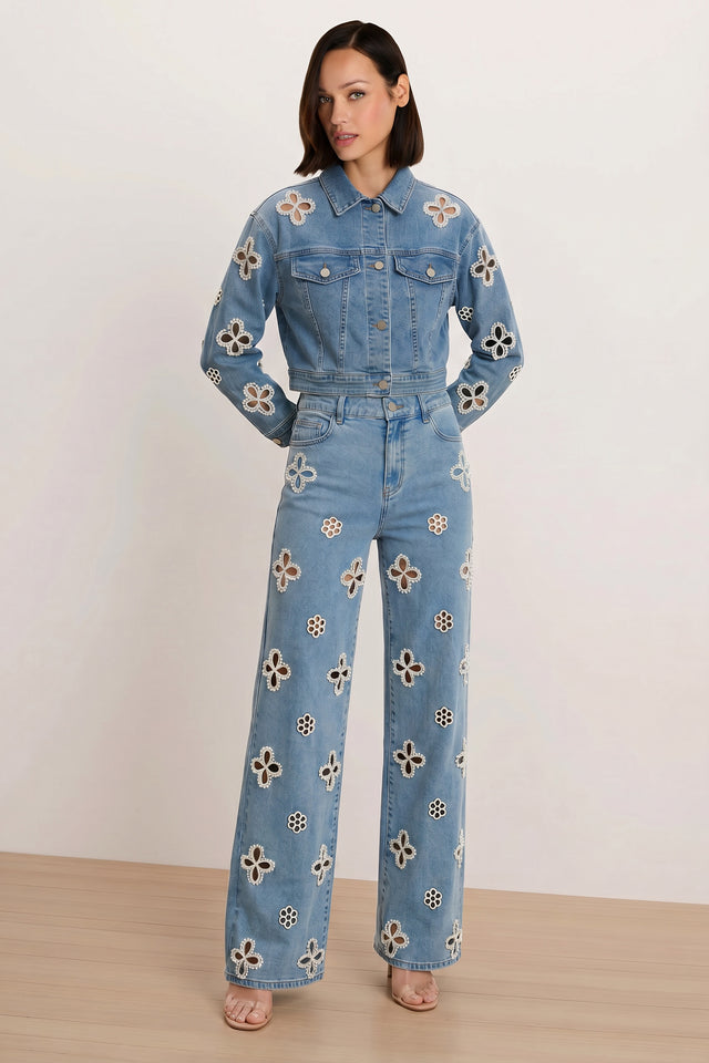 A woman with shoulder-length hair poses with hands in her pockets, wearing the Kaia Pearl High Rise Jean in medium blue and a light blue statement denim jacket, both featuring white and brown floral cutout patterns. She completes the look with nude sandals.