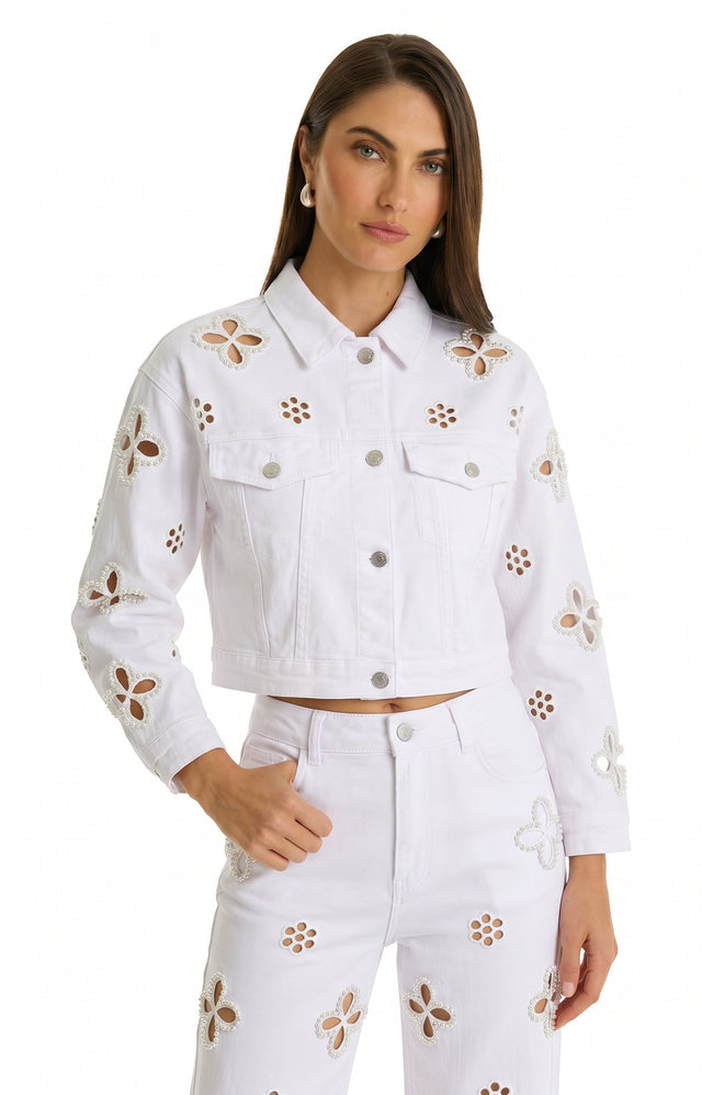 A woman with long brown hair and a neutral expression wears the Kaia Pearl Jean Jacket - White, featuring cutout floral patterns and delicate floral beadwork, paired with matching pants. She stands against a plain white background.