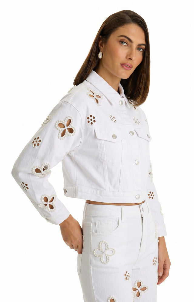A woman with straight brown hair and pearl earrings models the Kaia Pearl Jean Jacket in white, featuring floral beadwork, paired with matching high-waisted jeans. She poses against a white background with one hand by her side.