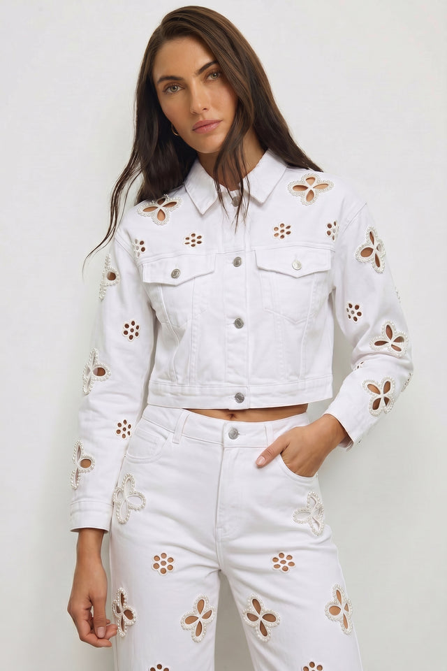 A woman wears the Kaia Pearl Jean Jacket - White with matching high-waisted jeans featuring cutout flower designs, standing against a plain light background for a modern, effortlessly stylish look.
