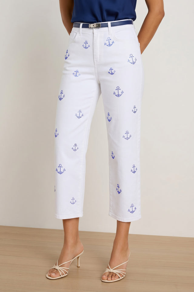 A person wears the Kelsey Cropped Jean - Crystal Anchors in white, paired with a navy belt, dark blue top, and strappy heeled sandals, standing on a wooden floor against a light background.