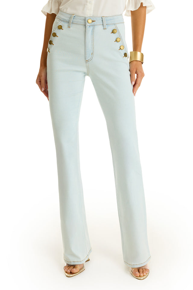 A person wearing the Allegra Bootcut Jean - Cloud, a white blouse, gold cuff bracelet, and open-toed heeled sandals poses against a white background.