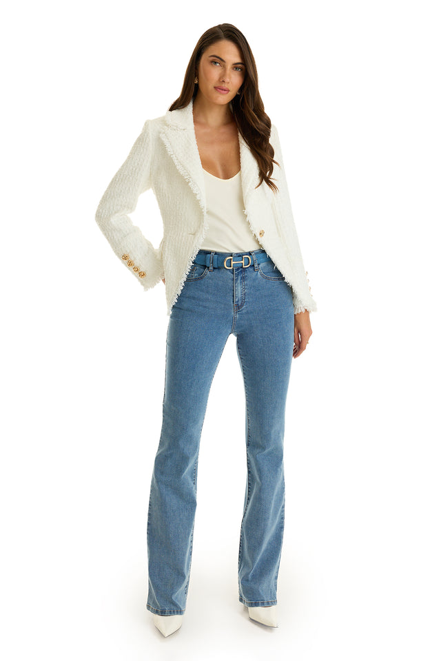 A woman stands confidently in the Giselle Single Breasted Tweed Blazer - White with gold buttons over a white top, paired with blue flared jeans, a belt, and white pointed-toe shoes against a plain white background.