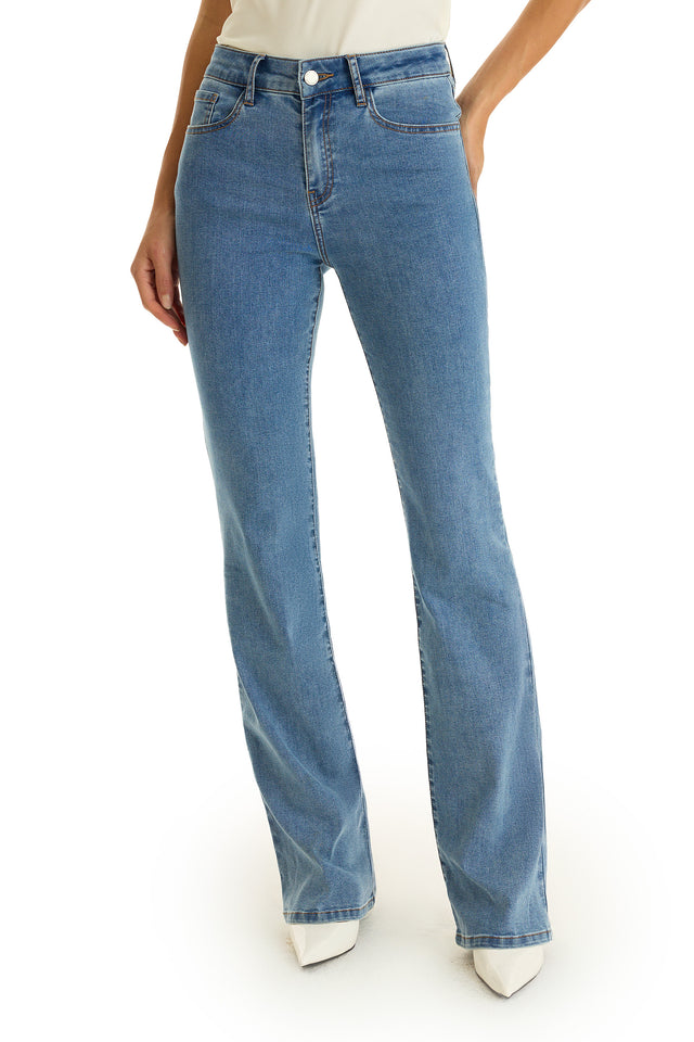 A person wears Aria Flare Jean in medium blue with white pointed-toe shoes, hands relaxed at their sides, photographed against a plain white background.