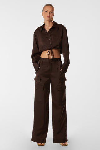A person stands in a stylish brown ensemble featuring the Milan Satin Button Up in espresso, completed with wide-leg cargo pants featuring tie-waist detailing. With hands in pockets and open-toe heels, they strike a pose against a plain white background.