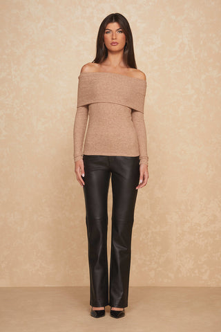 A woman stands against a textured beige background, wearing a praline Amora Ribbed Knit Top, highlighting its off-the-shoulder elegance. She pairs it with black leather pants and complements the look with her long, straight hair cascading down and black shoes, achieving a perfect balance of style and sophistication.