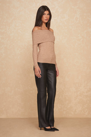A woman poses confidently against a beige textured wall, highlighting an off-the-shoulder Amora Ribbed Knit Top in Praline, combined with sleek black leather pants and pointed heels. Her long dark hair adds the finishing touch to this stylish and modern look.