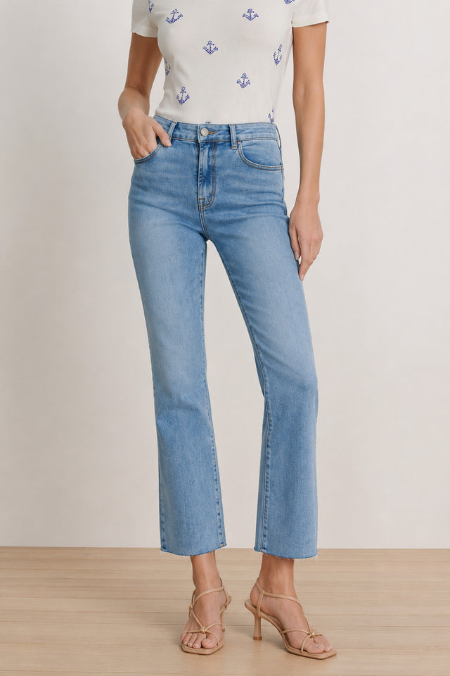 A woman stands against a plain background on a wooden floor, wearing the Nicolette Cropped Jean in medium blue with a straight-leg fit, paired with a white short-sleeve shirt featuring blue anchor prints and beige heeled sandals.