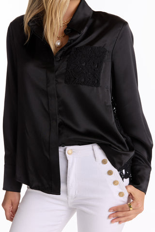 A woman wears the Amelia Silky Lace Blouse in black, featuring a lace pocket, paired with white high-waisted pants with gold front buttons. Only her torso and part of her head are visible.