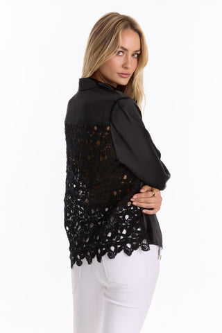 A woman models the Amelia Silky Lace Blouse - Black (Preorder), featuring an open lace back and long sleeves, paired with white pants as she stands slightly turned to the side against a white background.