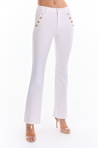 A woman stands against a white background wearing the Allegra Bootcut Jean - White (Preorder) with gold front buttons, paired with clear high-heeled sandals.