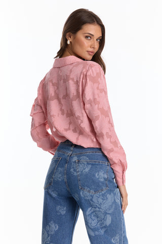 A woman, facing away and glancing over her shoulder, wears the Stelle Button Up - Rose, a light pink blouse with subtle floral lace details, paired with blue jeans. The blouse’s lightweight fabric stands out against the white background.