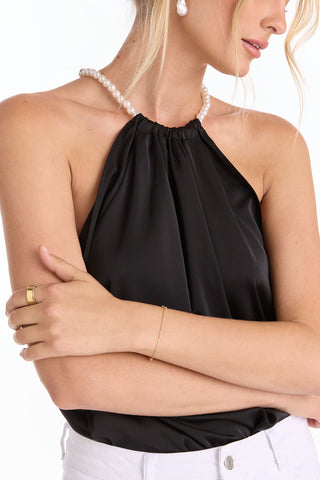 A woman with light hair and white pants poses against a white background, wearing the Mika Silky Halter Top - Black (Preorder), styled with a pearl necklace, gold ring, and thin gold bracelet for a statement look.