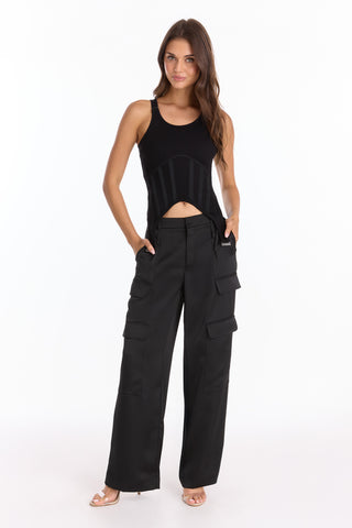 A woman with long brown hair stands against a white background, smiling slightly as she wears the Kiara Garter Tank - Black and black wide-leg cargo pants with multiple pockets, open-toed heels, and her hands in her pockets.