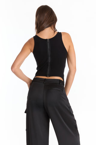 A woman stands facing away in the Kiara Garter Tank - Black, featuring rib fabric and a visible back zipper, paired with high-waisted black wide-leg pants. Her right hand rests on her hip against a white background.
