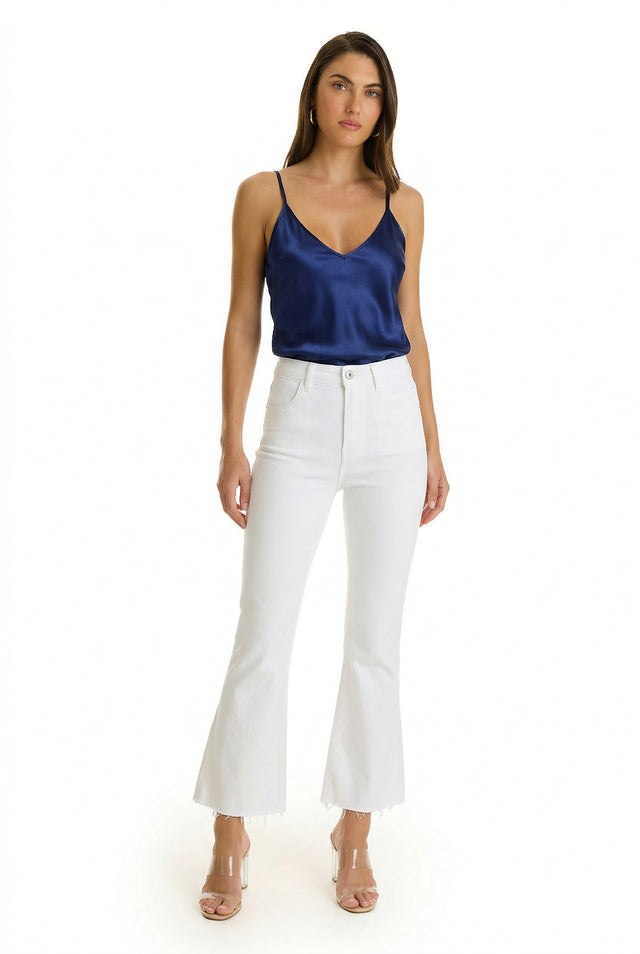 A woman poses against a white background in a navy Yara Silk Camisole with a deep v-neck, high-waisted white flare jeans with frayed hems, clear strap heels, and hoop earrings.