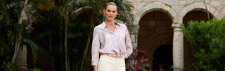 A woman stands outdoors in front of stone arches and lush greenery, wearing a light pink patterned blouse tucked into beige pants with a matching belt.