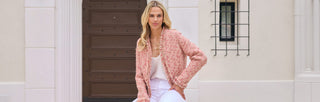 A woman with blonde hair, wearing a pink textured jacket and white pants, sits on a white bench in front of a large, arched wooden door with cream-colored walls and brick steps.